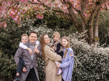 Family of five smiles in front of cherry blossoms